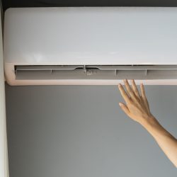 Woman wants to catch stream of warm air stretching hand to device slot at home. Female turns on air conditioner hanging on grey wall to warm up apartment to feel better